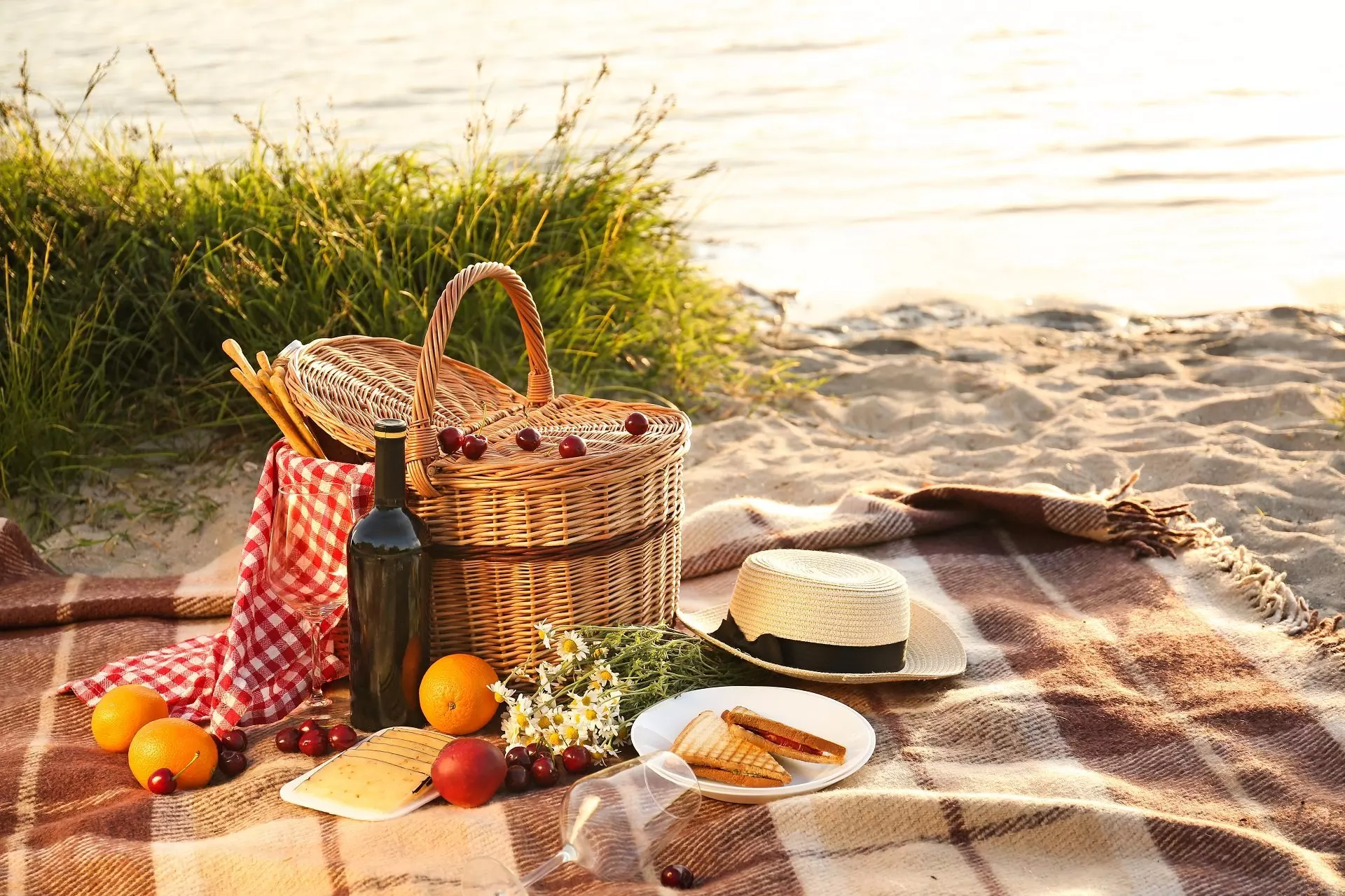 Pepco picnic set on beach blanket with wicker basket, straw hat, wine bottle, fruits, cheese and sandwiches by the water.