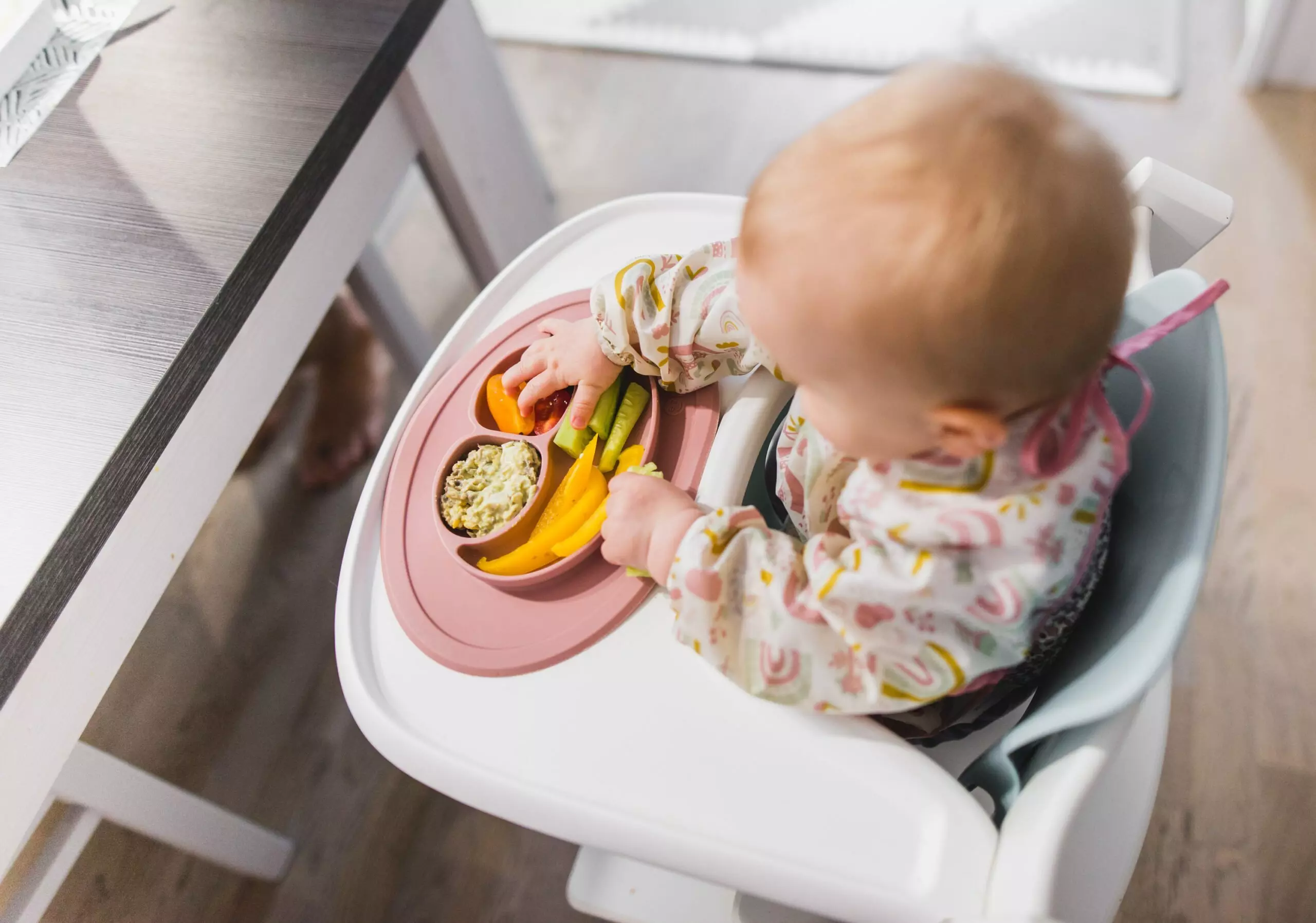 Baby in highchair eating from Pepco pink sectioned plate with vegetables and food in compartments.