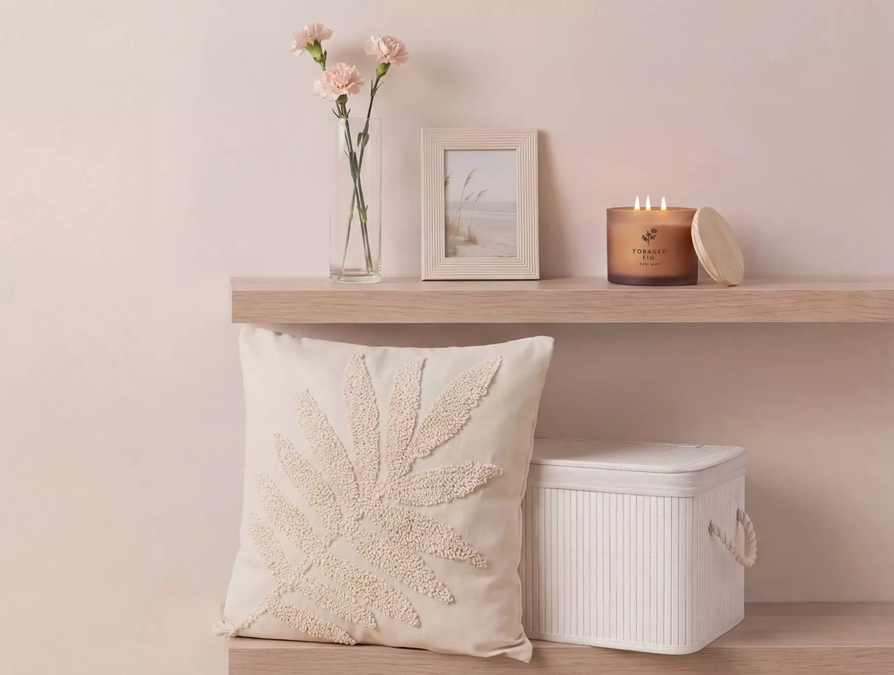 Beżowa aranżacja wnętrza Pepco z haftowaną poduszką, świeczką, wazonem różowych kwiatów i ramką na zdjęcia na drewnianej półce.
