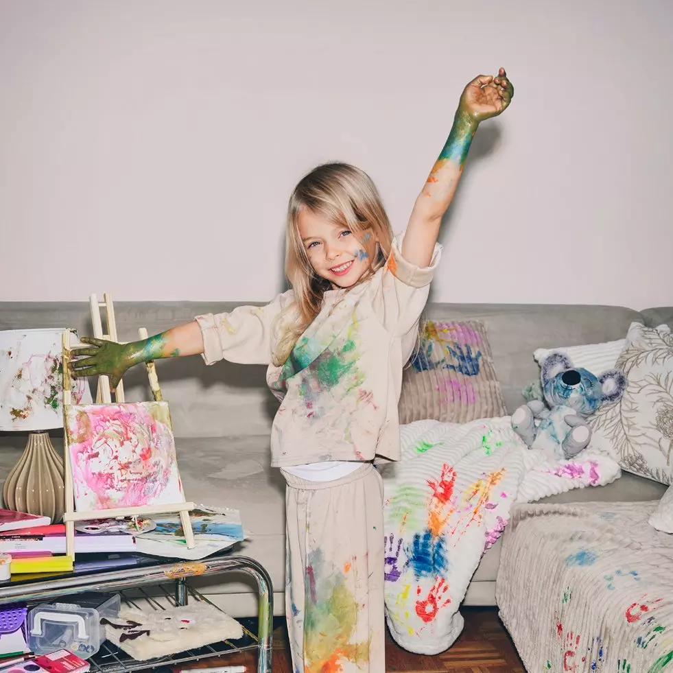 Child with paint-covered hands and clothes joyfully painting, standing next to art easel on sofa with colorful handprints and teddy bear nearby.