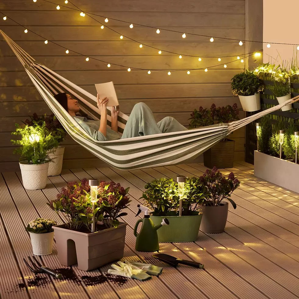 Person relaxing in striped hammock reading a book on wooden deck with string lights and potted plants.