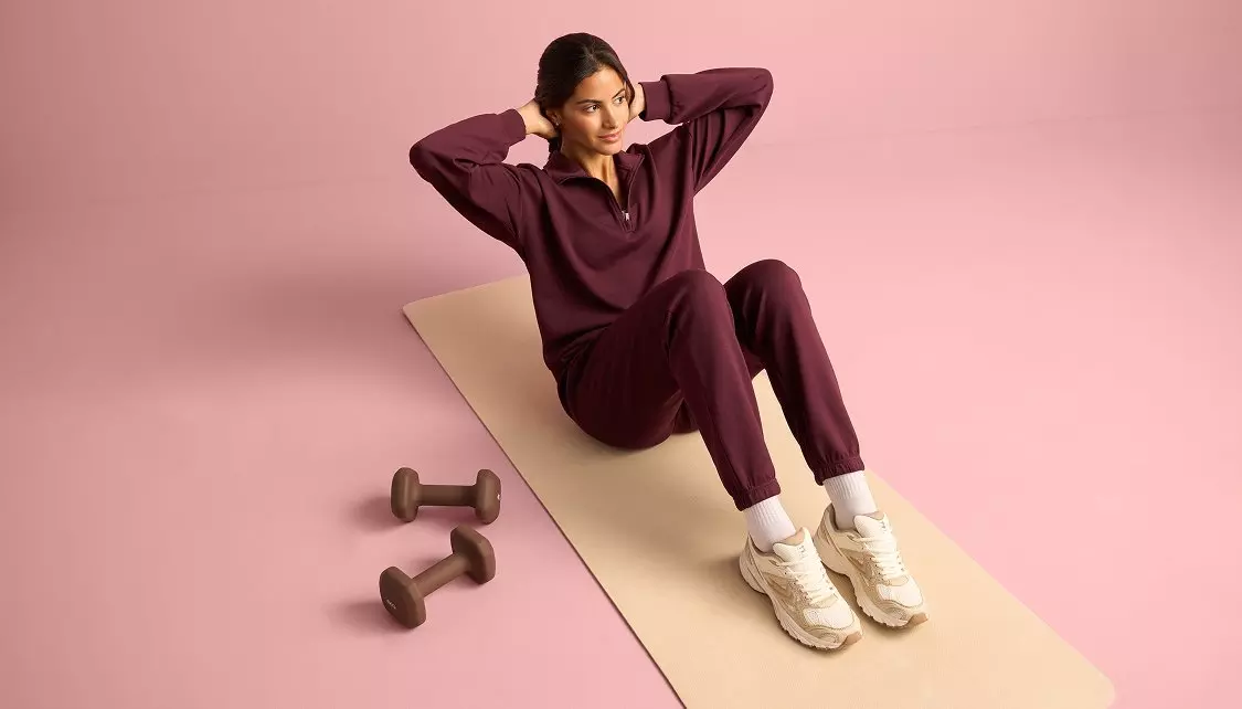 Pepco woman wearing burgundy tracksuit sitting on exercise mat with dumbbells nearby.