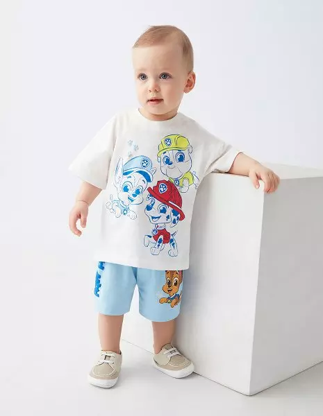 Chłopiec w białej koszulce z postaciami Psi Patrol i niebieskich szortach stoi przy białym sześcianie.