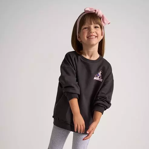 Niña sonriente lleva sudadera negra Pepco con detalle bordado y lazo rosa en el pelo.
