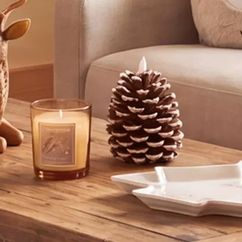 Vela aromática Pepco y piña decorativa sobre mesa de madera con sofá beige al fondo.