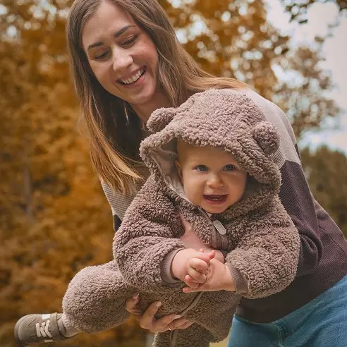 Mamma sorridente tiene in braccio un bambino che indossa una giacca Pepco in pile marrone con cappuccio.