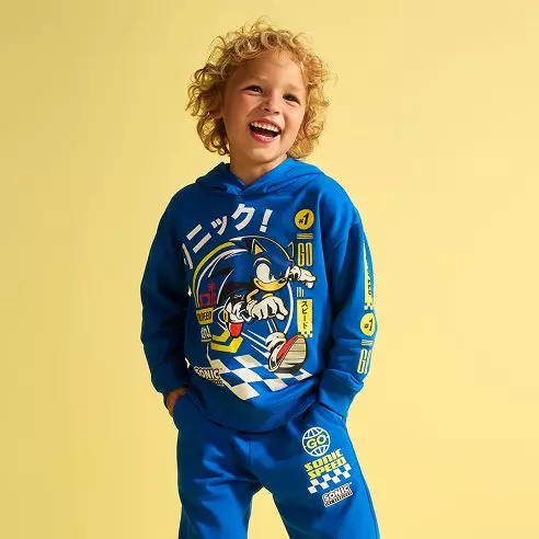 Dieťa v modrej mikine a nohaviciach Pepco so Sonic the Hedgehog potlačou sa usmieva.