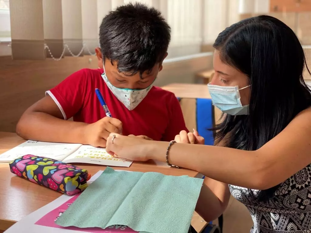 O femeie ajută un băiat care poartă mască să scrie la o masă cu caiete și un penar colorat.