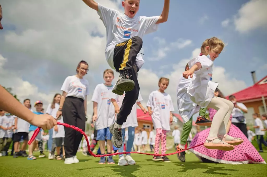 Copii sărind peste frânghie într-o activitate în aer liber, purtând tricouri albe.