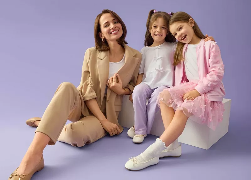 Kobieta w beżowym garniturze i dwie dziewczynki w pastelowych strojach uśmiechają się do kamery.