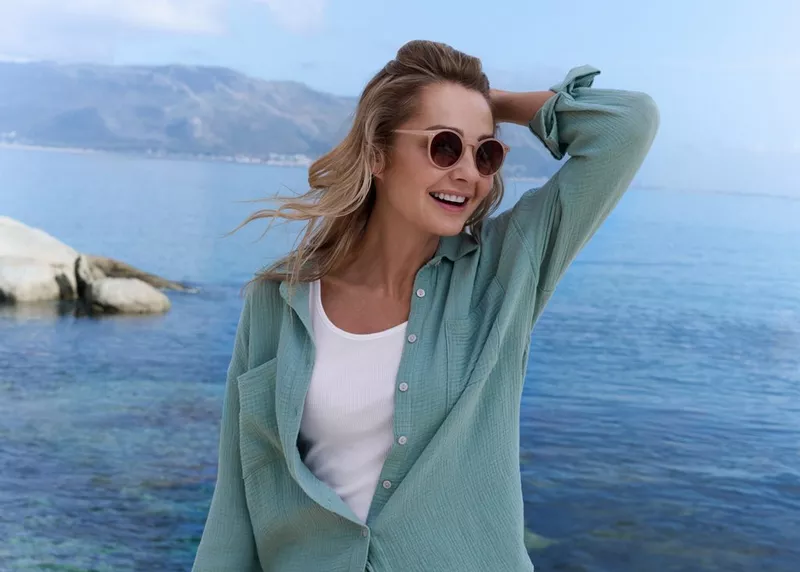 Kobieta w miętowej koszuli i okularach przeciwsłonecznych uśmiecha się nad brzegiem morza.