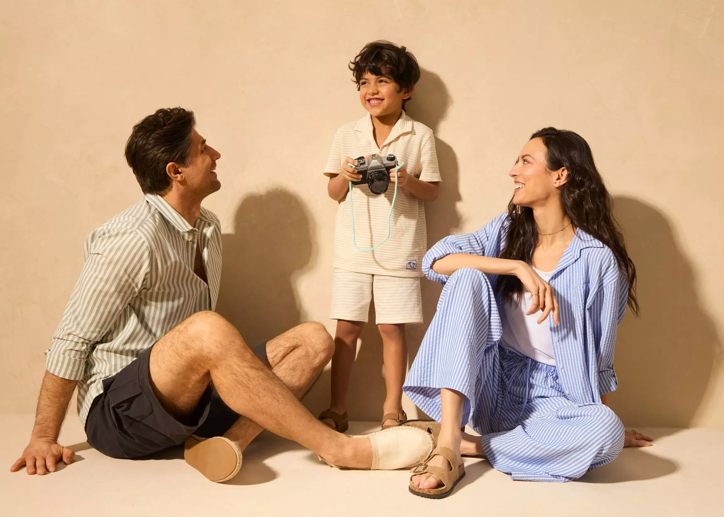 Copil ținând un aparat foto în timp ce stă între părinții săi zâmbitori, purtând pijamale în dungi.