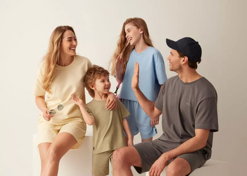 Familia sonriendo vistiendo conjuntos de camisetas y pantalones cortos en tonos beige, azul y gris.