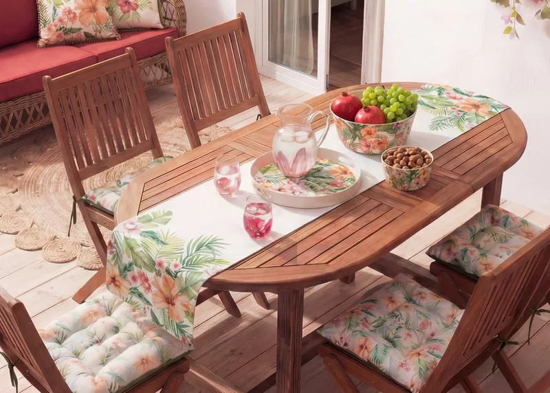 Mesa de madera ovalada con sillas, decorada con camino floral, frutas frescas y vajilla tropical.