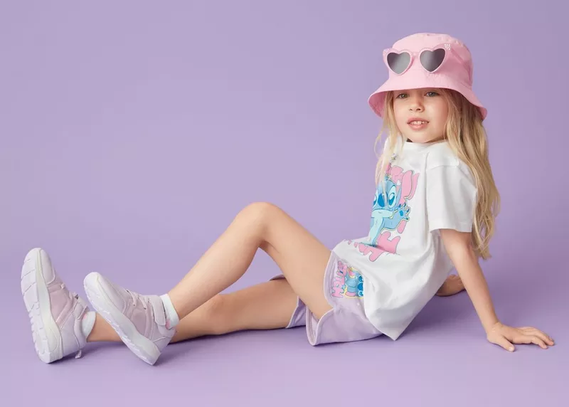 Girl wearing a pink bucket hat, white character print t-shirt, lilac shorts and trainers, sitting.