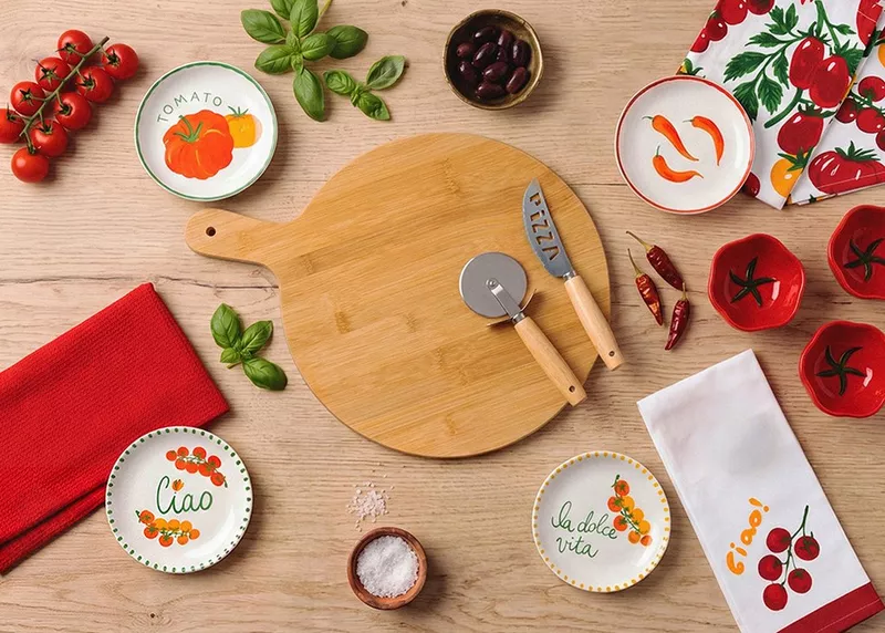 Pepco bamboo chopping board with pizza cutter surrounded by Italian-themed plates, fresh tomatoes, basil and chillies.