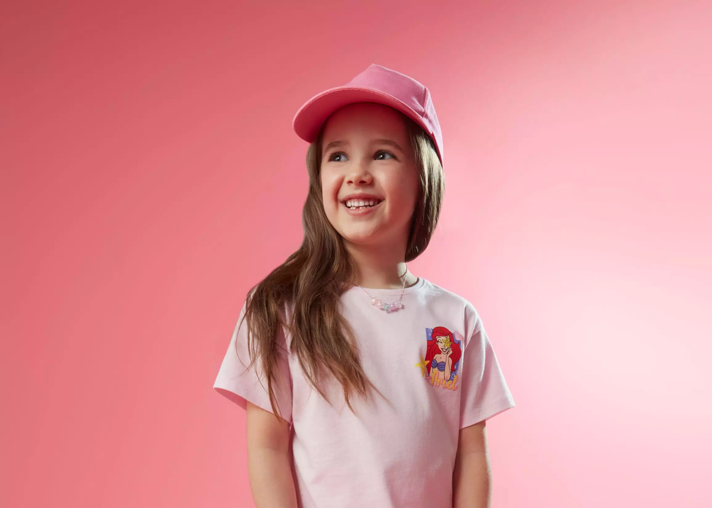Fată zâmbitoare purtând șapcă roz și tricou Pepco cu imprimeu Ariel Disney.