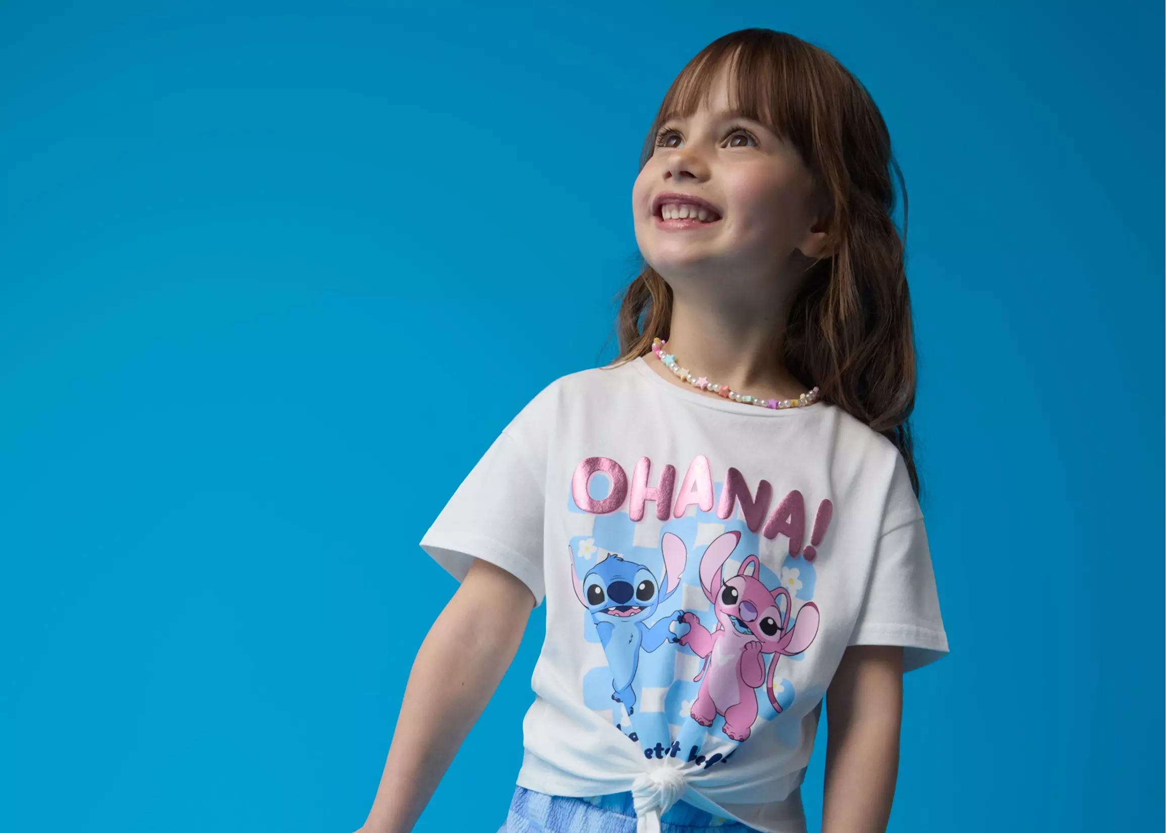 Niña sonriendo con camiseta blanca de Pepco con personajes de Lilo & Stitch y texto OHANA.