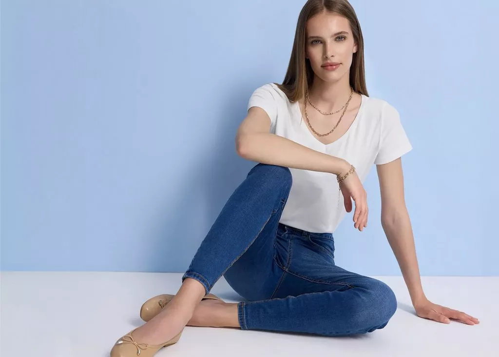 Mulher sentada usando t-shirt branca Pepco, calças de ganga azuis e sapatilhas bege com colares dourados.