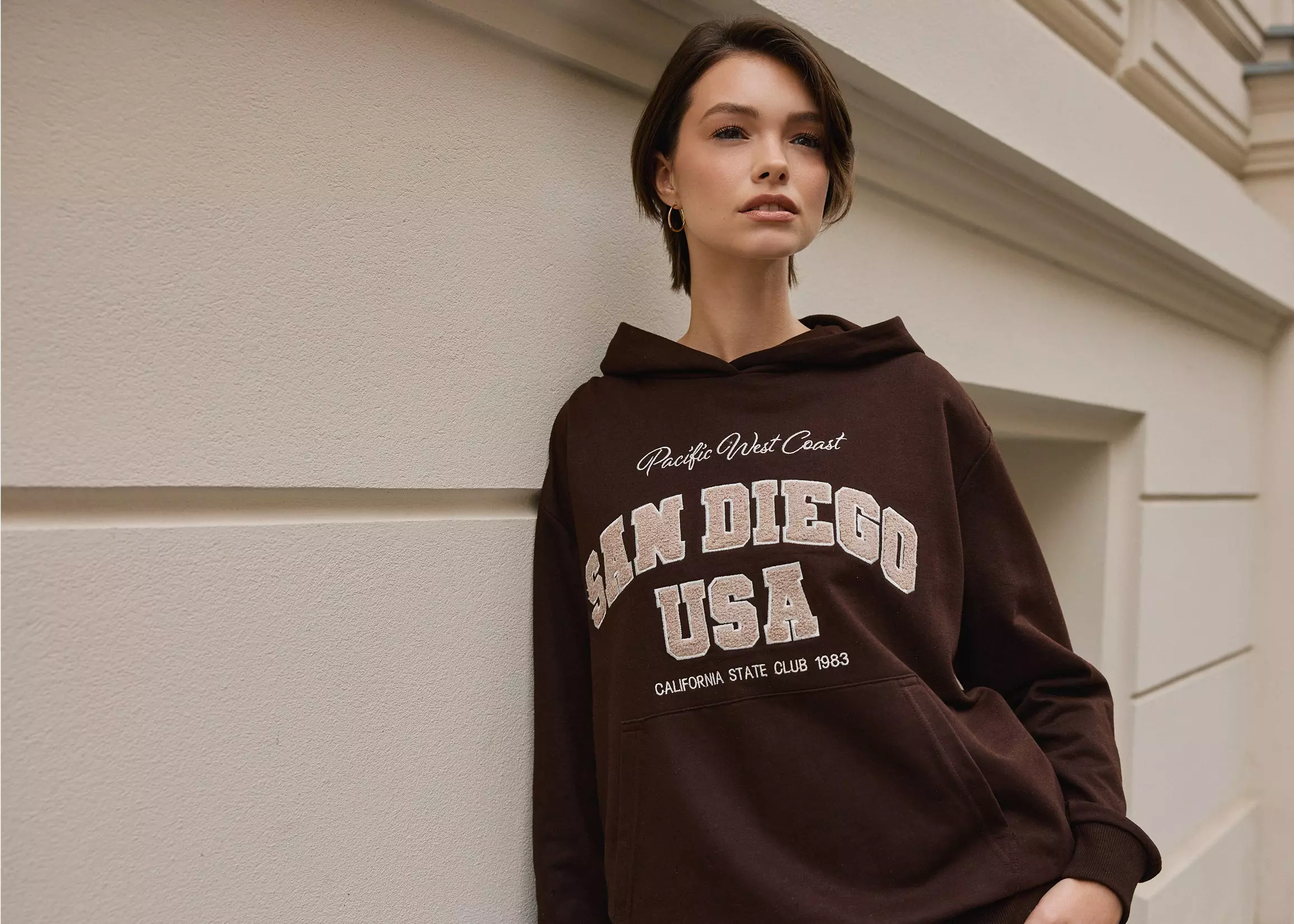 Žena v hnedej mikine Pepco s nápisom San Diego USA a kapucňou.