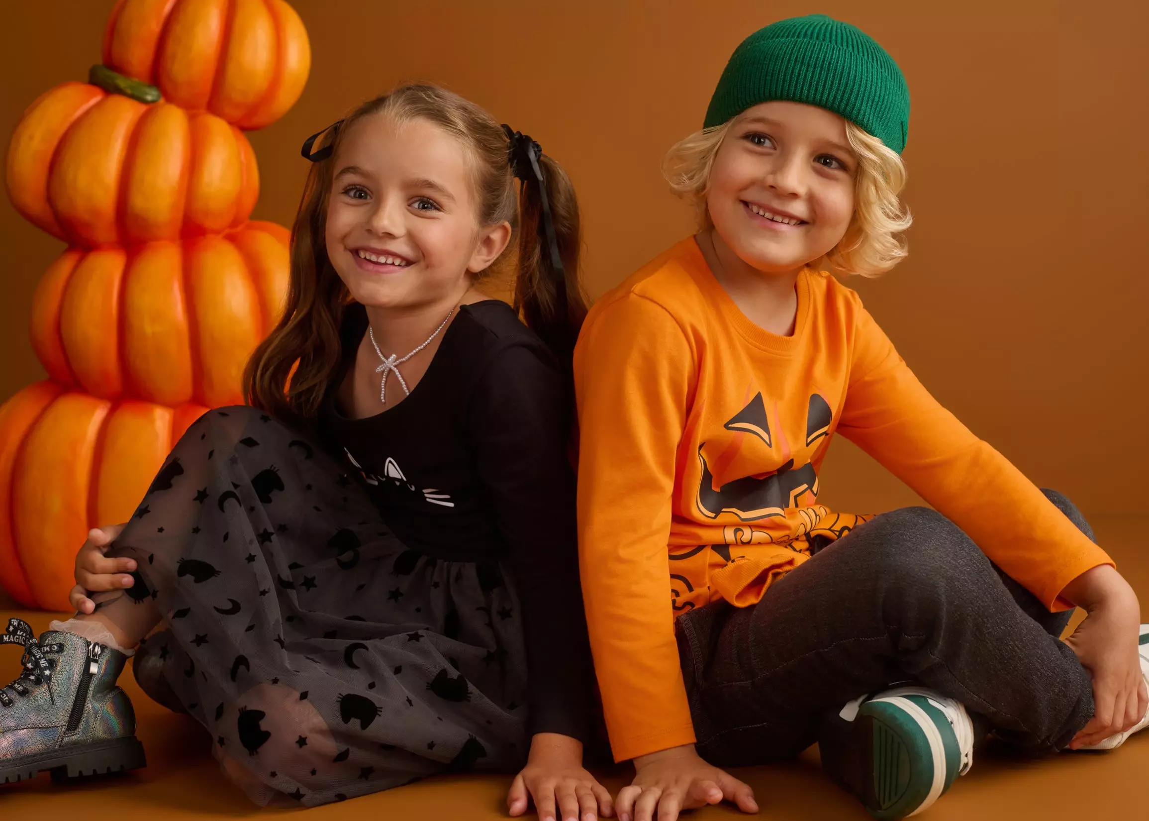 Due bambini sorridenti in abbigliamento Pepco a tema Halloween con decorazioni di zucche arancioni.