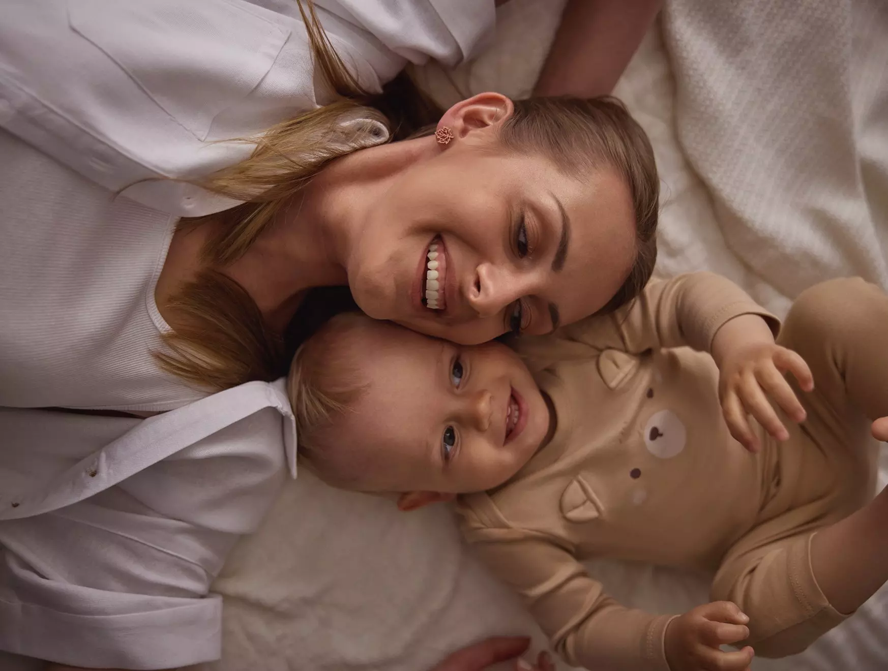 Pepco mother and baby lying together on bed, both smiling at camera wearing matching beige clothing.