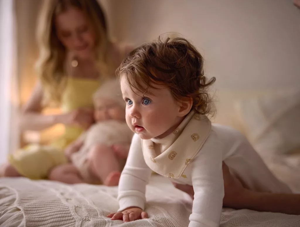 Bambino che gattona indossando abbigliamento comodo Pepco mentre la mamma gioca con un neonato sullo sfondo.