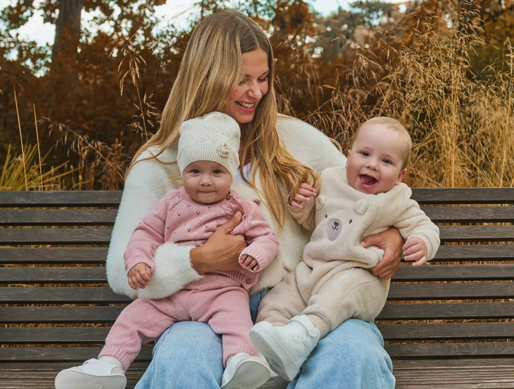 Smiling mother sitting on a bench holding two babies wearing Pepco knitwear in pink and cream during autumn.