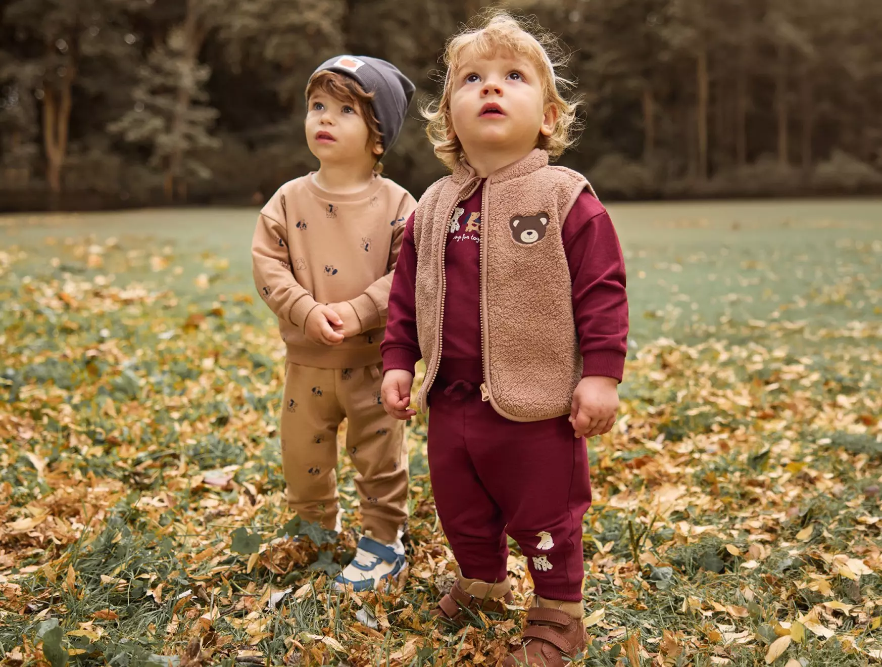 Two young children in Pepco autumn outfits standing amongst fallen leaves, looking upward in a woodland setting.