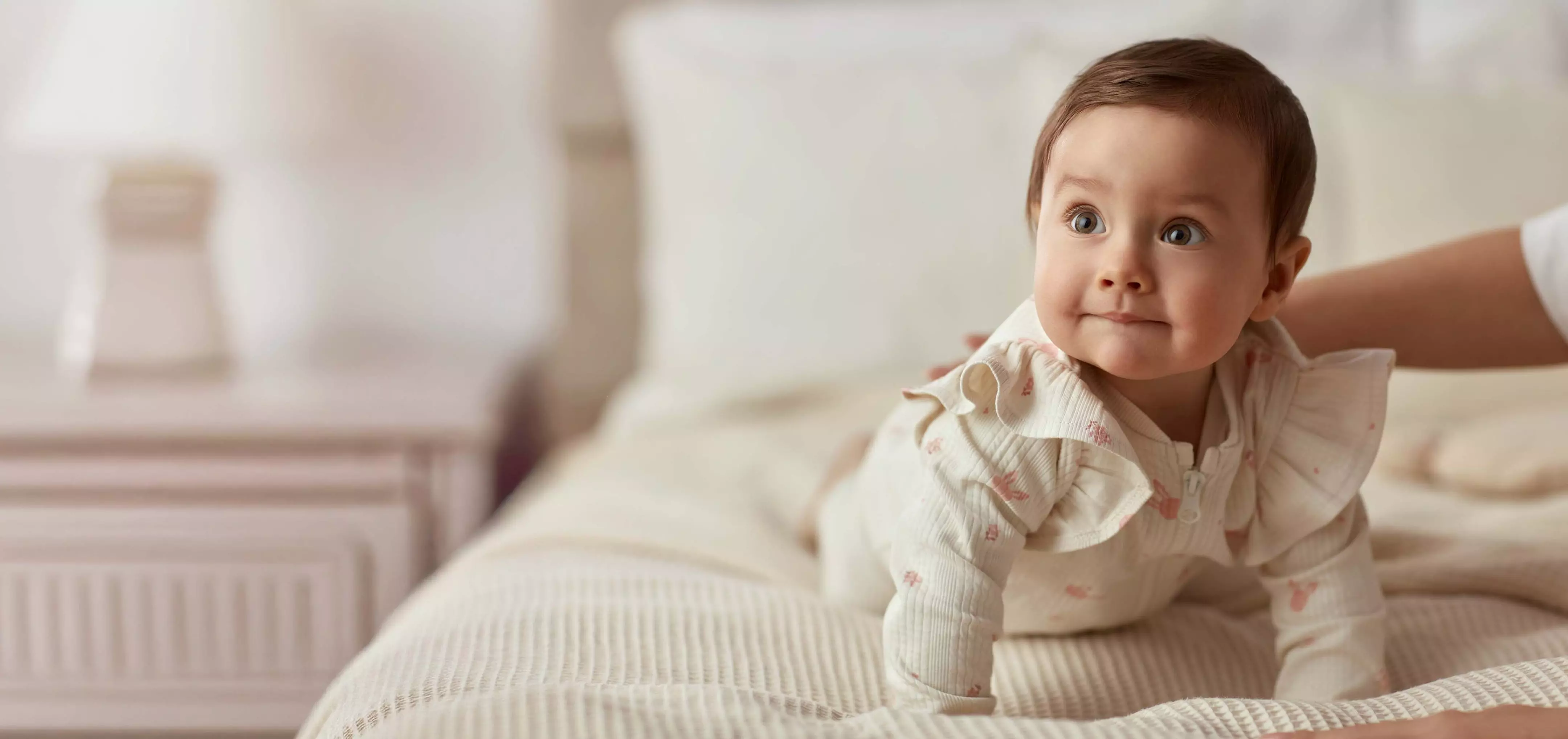 Baby wearing cream-colored outfit with subtle pattern sitting on textured bedding in bright room.