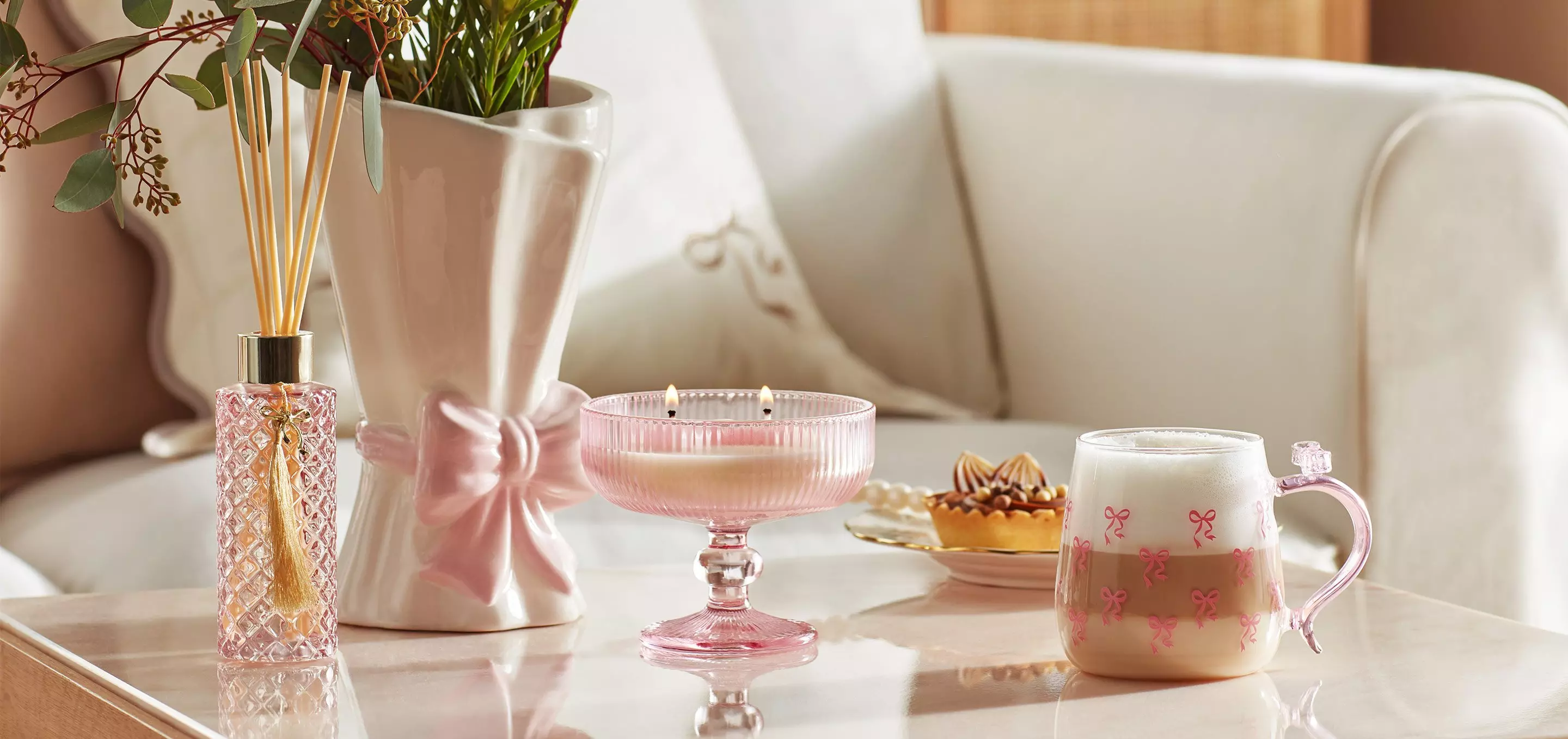 Eleganckie różowe akcesoria domowe Pepco: dyfuzor zapachowy, wazon z kokardą, świeca i kubek na stoliku kawowym.