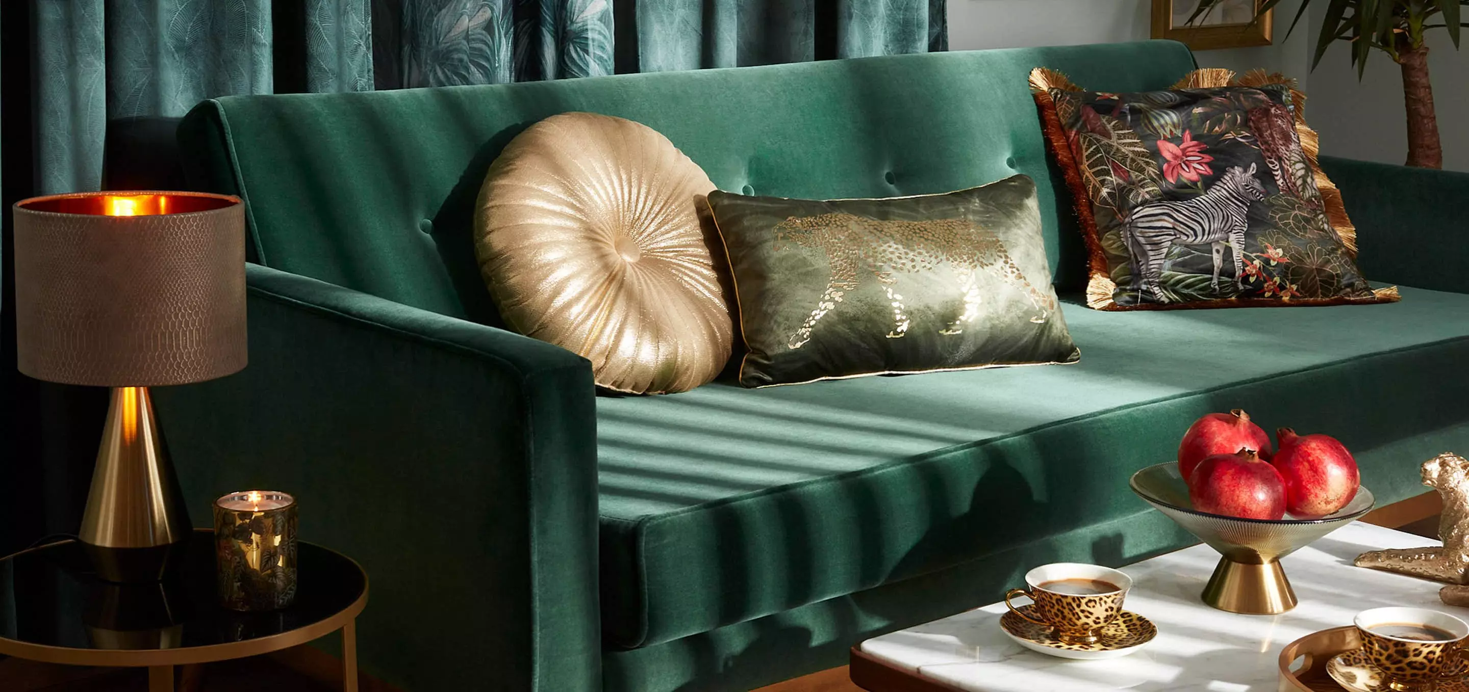 Elegancka zielona sofa Pepco z ozdobnymi poduszkami, lampą stołową i misą owoców na stoliku kawowym.