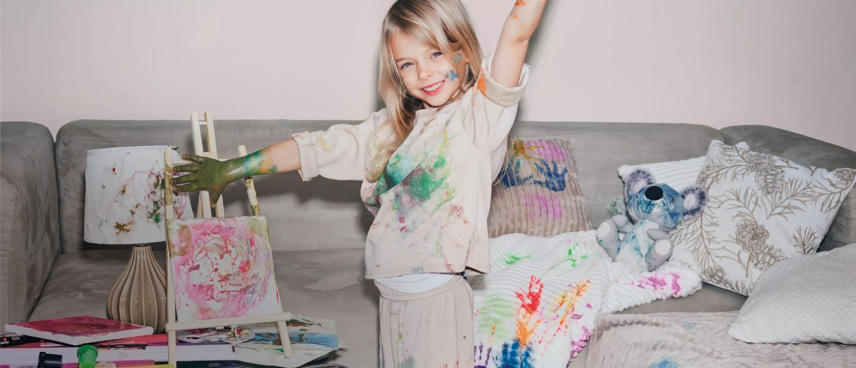 Dziecko w poplamionych farbą ubraniach malujące na sztaludze, z kolorowymi śladami farby na rękach i otoczeniu.