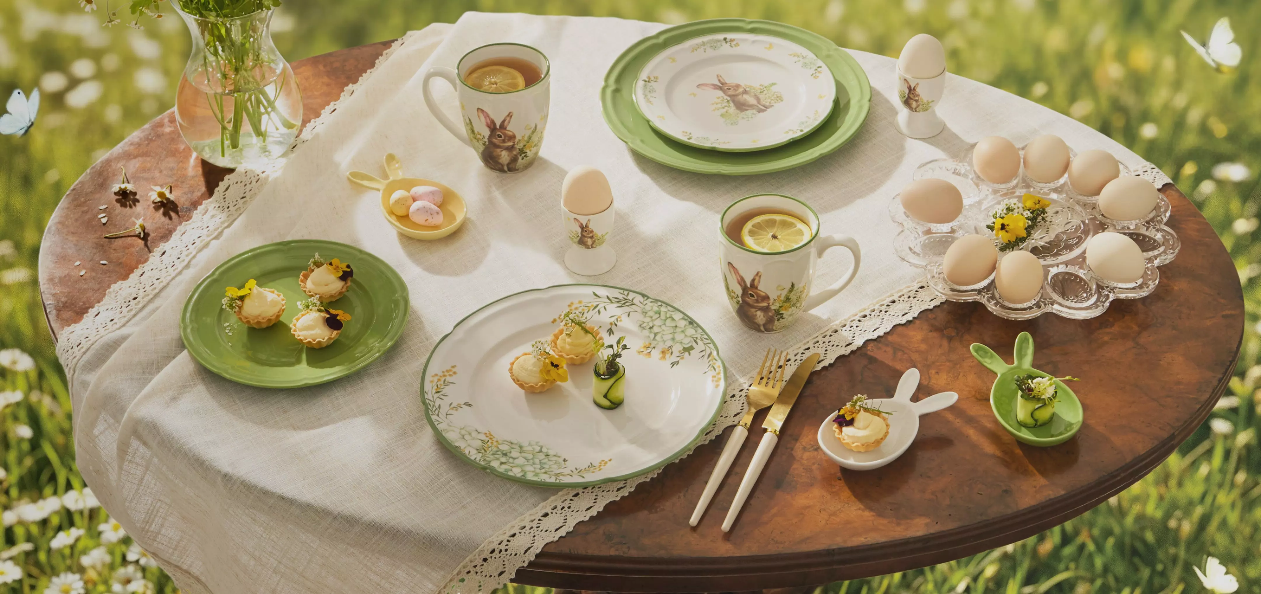 Easter table setting with bunny-themed tableware, tea cups, small pastries, and eggs arranged on a wooden table outdoors.