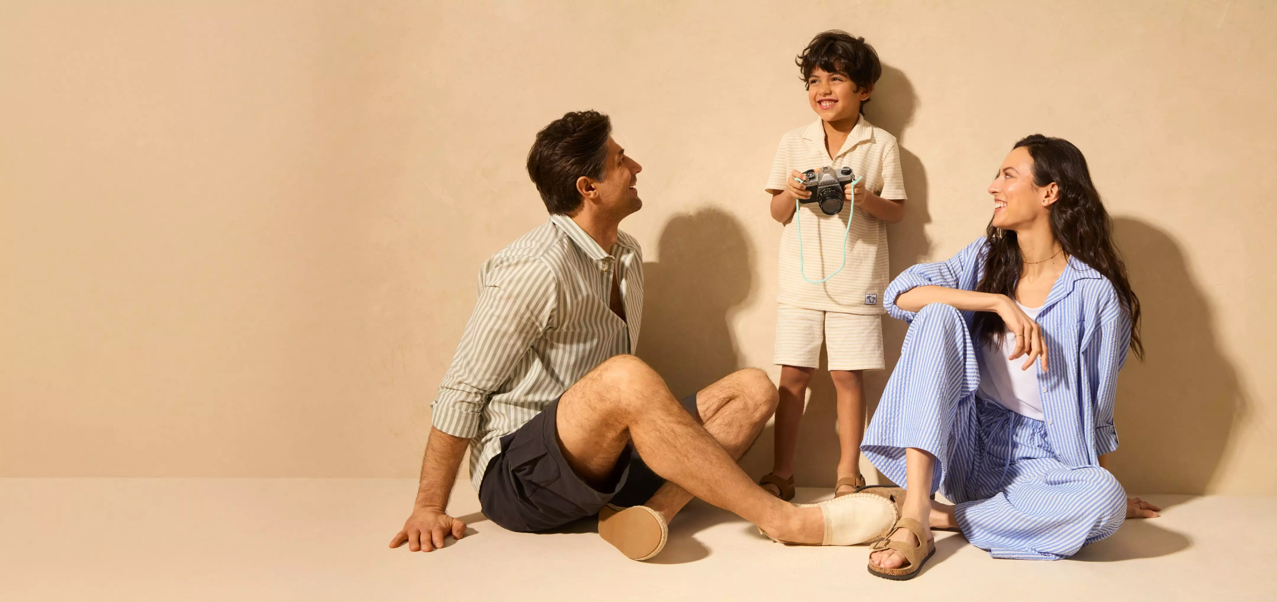 Familie relaxată cu părinți și copil ținând un aparat foto, toți purtând haine casual în dungi.