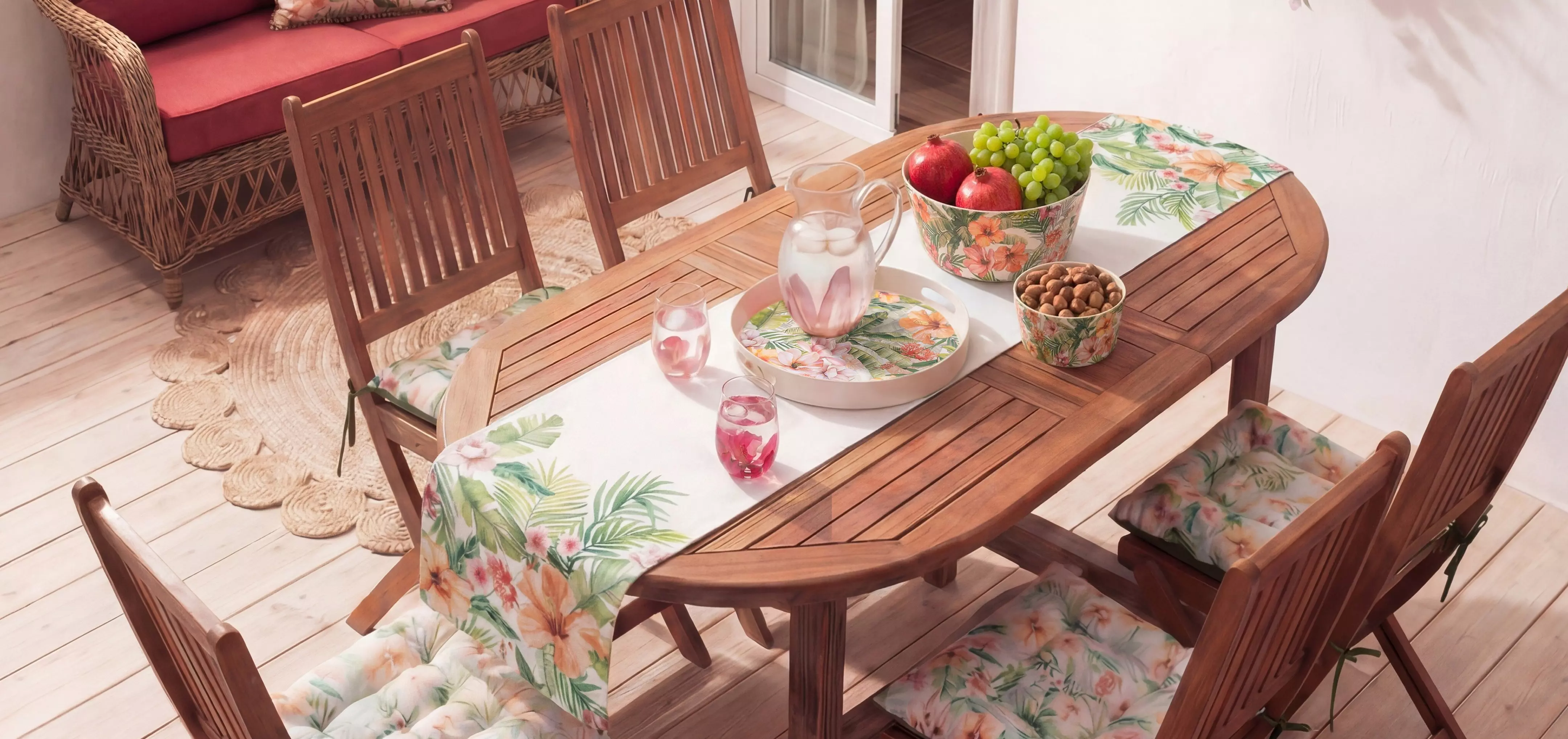 Mesa de madera ovalada con cuatro sillas, decorada con vajilla tropical, frutero y copas con bebida.