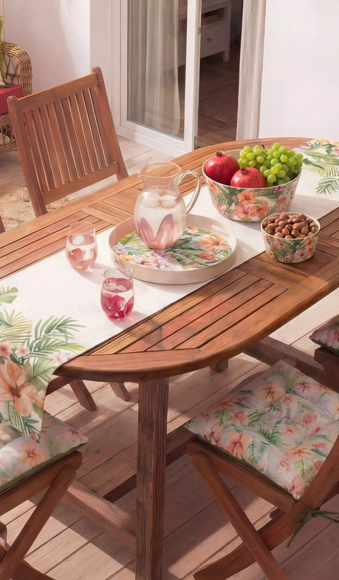 Mesa de madera para exteriores con vajilla floral, cuenco de frutas, jarra y cojines estampados en terraza.