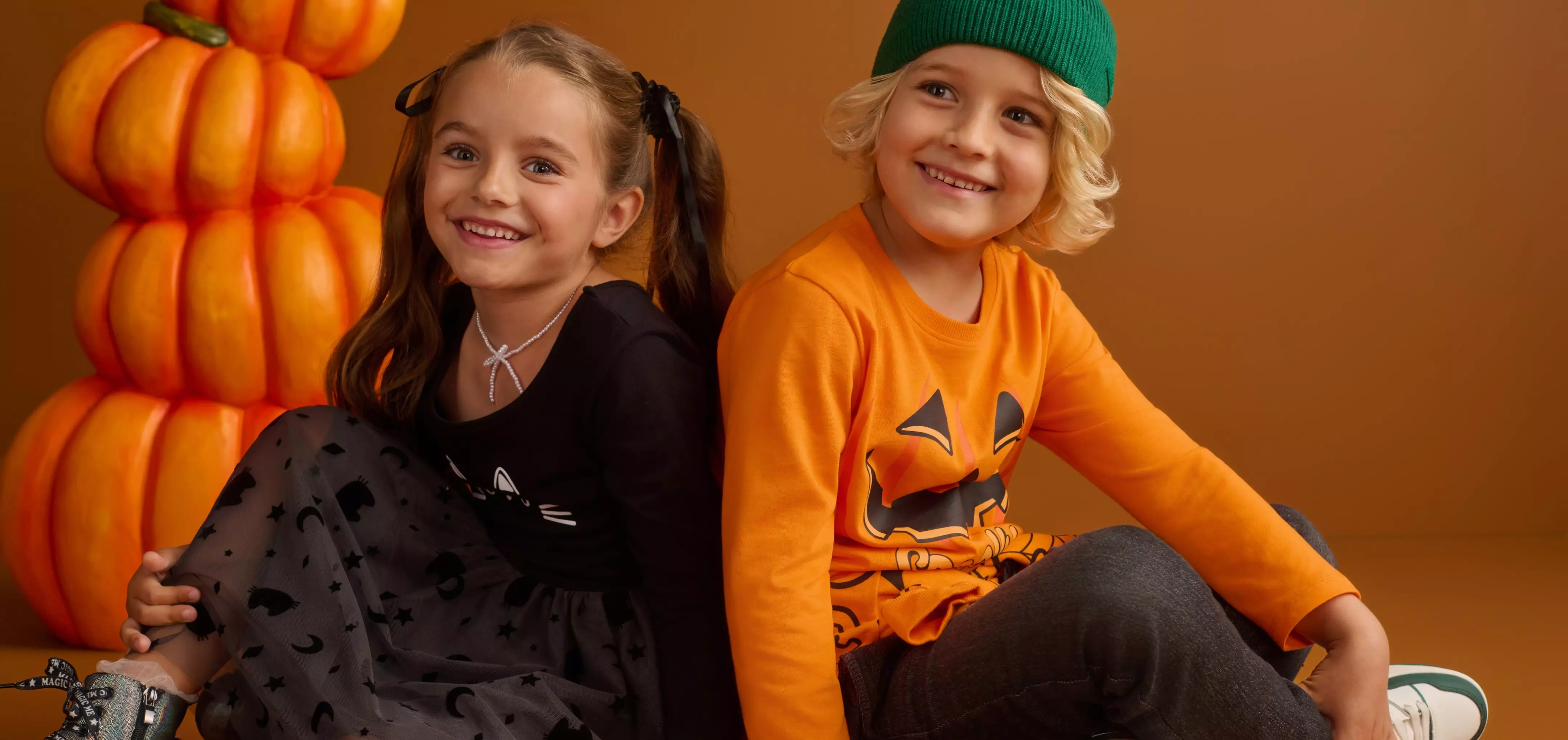 Due bambini sorridenti in abbigliamento Pepco a tema Halloween con decorazioni di zucche arancioni.