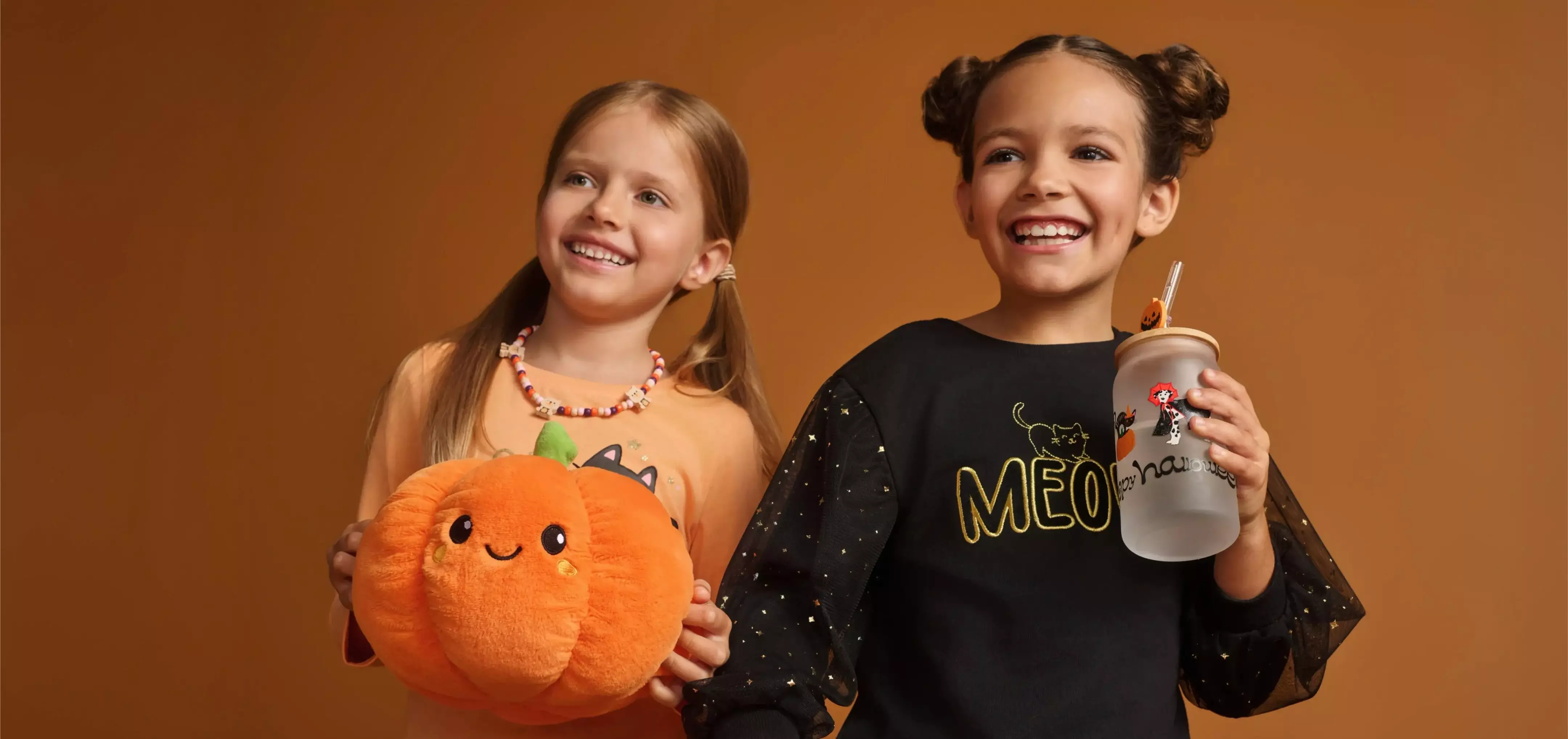 Dwie uśmiechnięte dziewczynki Pepco w strojach Halloween, jedna trzyma pluszową dynię, druga kubek.