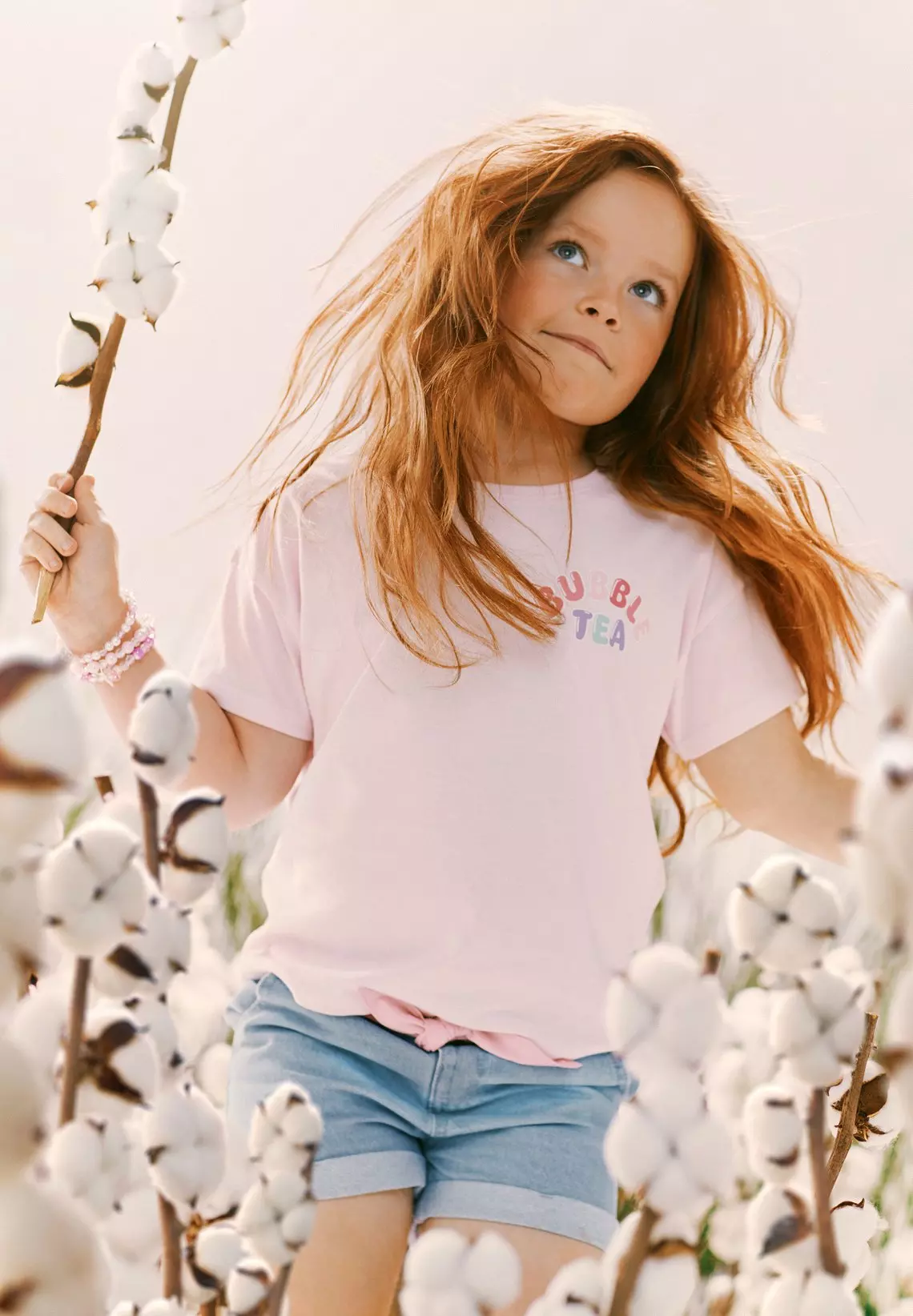 Niña con cabello pelirrojo sostiene una rama de algodón, viste camiseta rosa y shorts vaqueros.