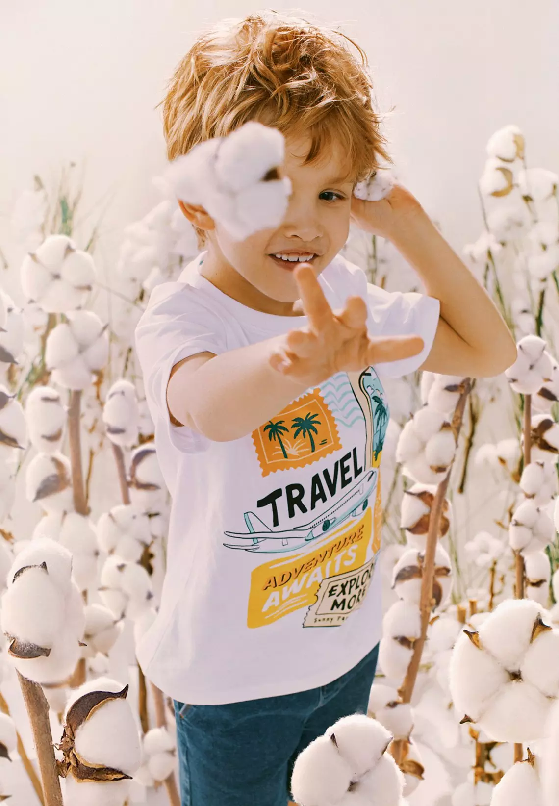 Niño sonriente con camiseta blanca estampada con "TRAVEL" en un campo de algodón.