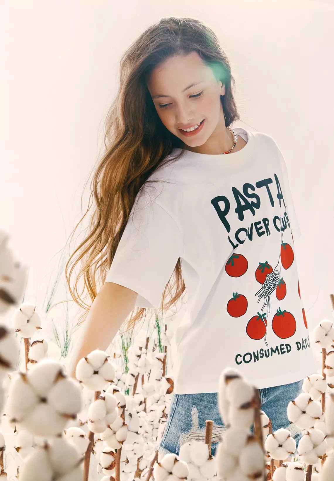 Mujer sonriente con camiseta blanca con texto PASTA LOVER CLUB y gráficos de tomates en campo de algodón.