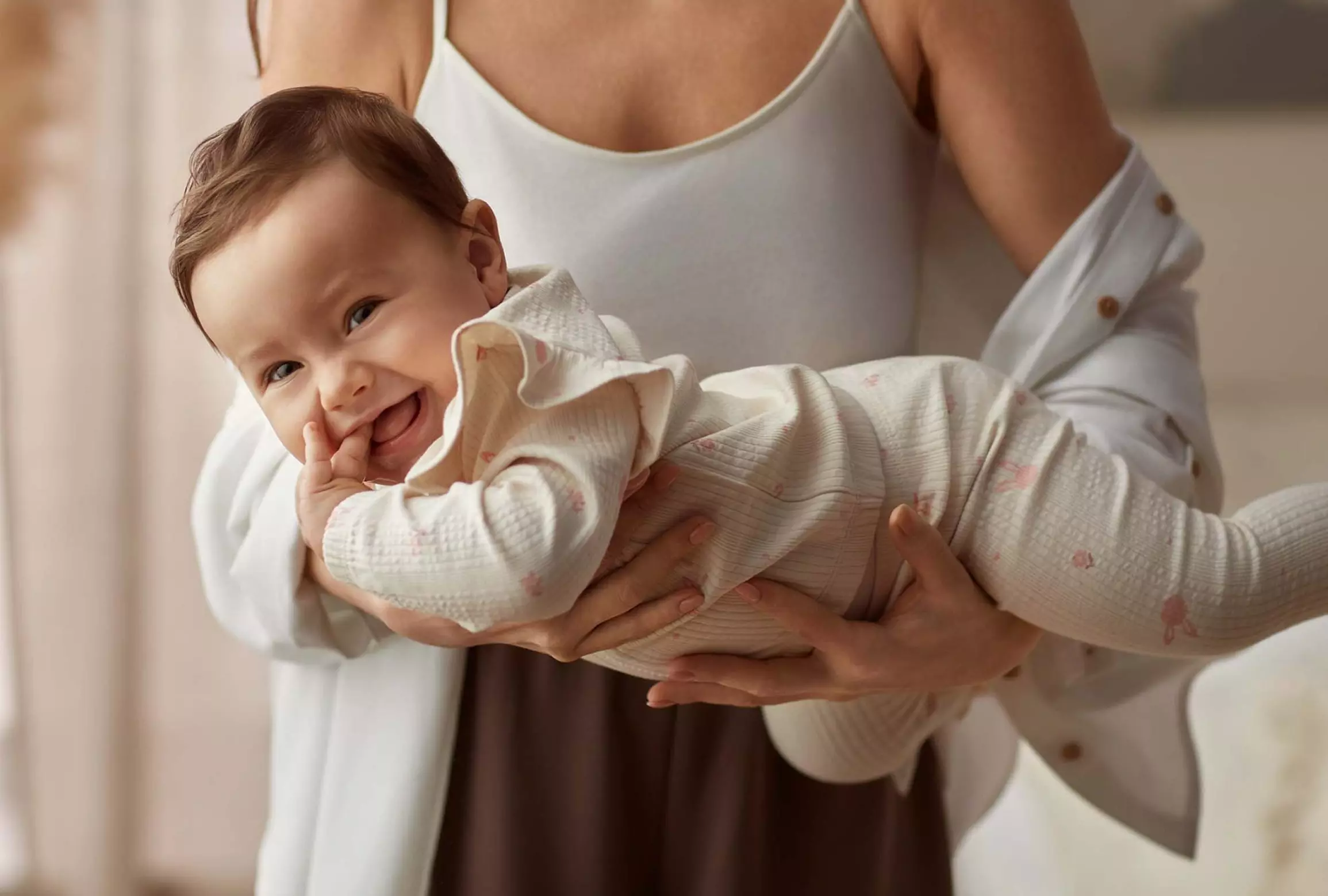 Uśmiechnięte niemowlę w białym ubranku z delikatnym wzorem, trzymane na rękach przez opiekuna w jasnej odzieży.