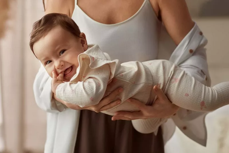 Uśmiechnięte niemowlę w białym ubranku z delikatnym wzorem, trzymane na rękach przez opiekuna w jasnej odzieży.