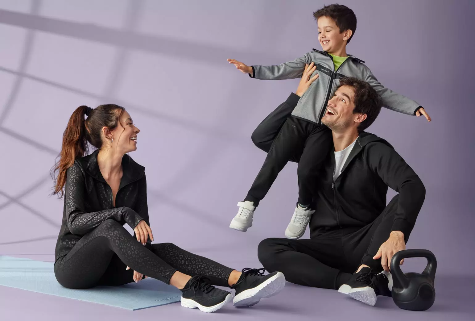 Rodzina w sportowych strojach Pepco podczas zabawy - dziecko na ramionach dorosłego, kobieta siedzi na macie fitness obok kettla.