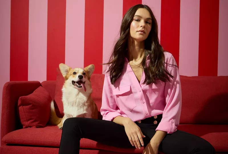 Pepco pink button-up shirt modeled with black pants, sitting on red sofa with corgi dog against striped background.