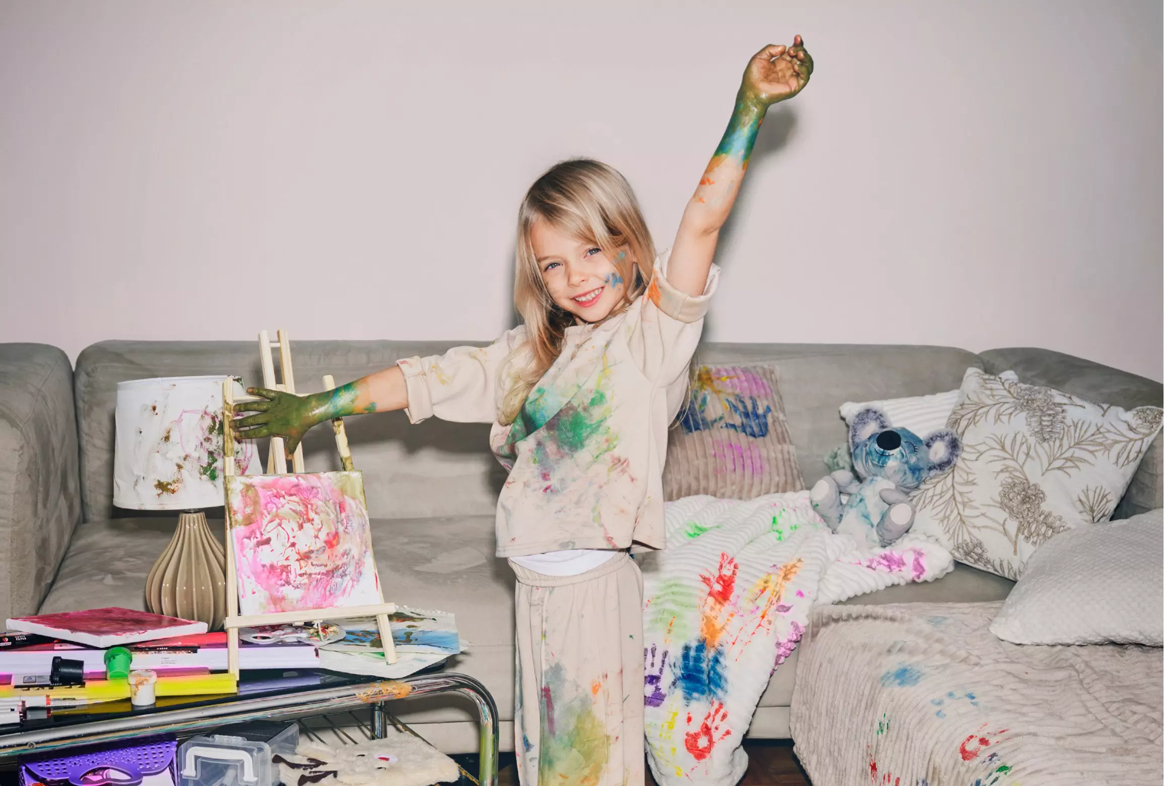 Child with paint-covered arms standing on sofa next to art supplies and colorful paintings.