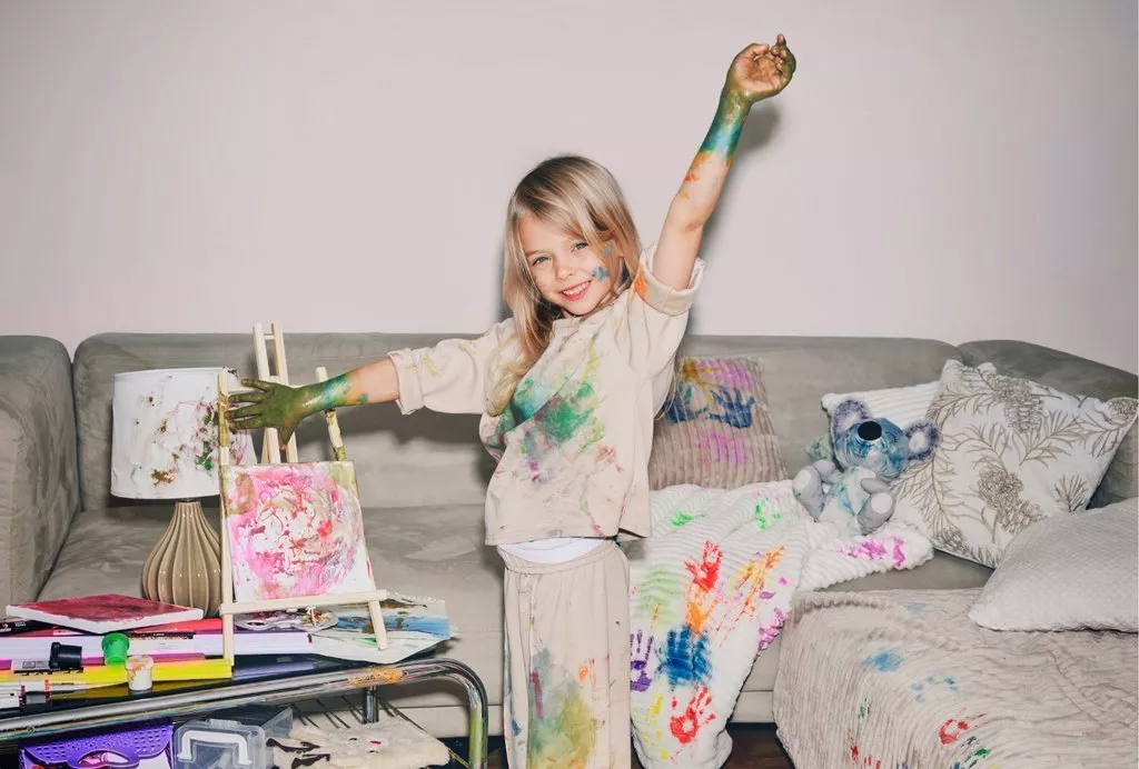 Child with paint-covered arms standing on sofa next to art supplies and colorful paintings.
