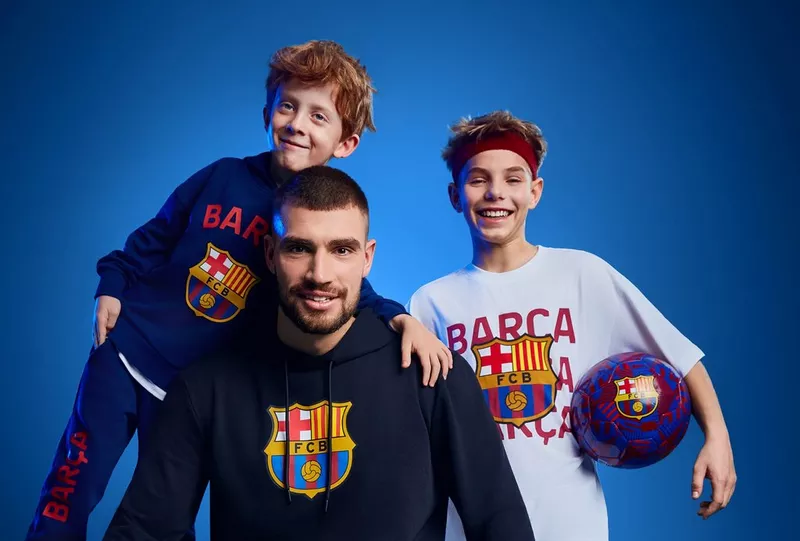 Trzech fanów FC Barcelona w oficjalnej odzieży klubowej, jeden trzyma piłkę z herbem klubu.