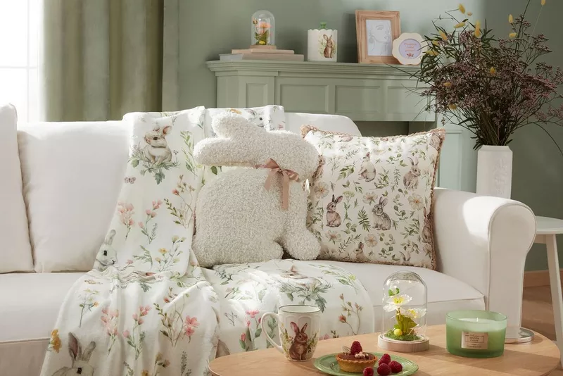 Biała sofa z poduszkami w kwiatowe wzory i pluszowym królikiem. Na stoliku kubek, tartaletka z malinami i świeca.